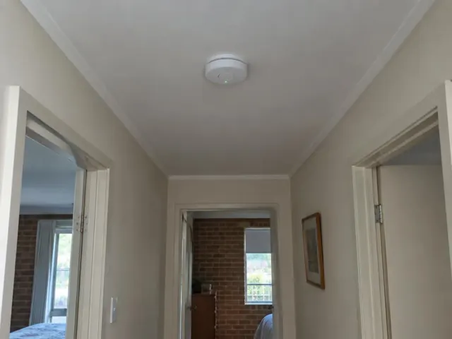Hallway ceiling smoke alarm above a beige interior corridor with doorways and brick wall glimpses in Kincumber